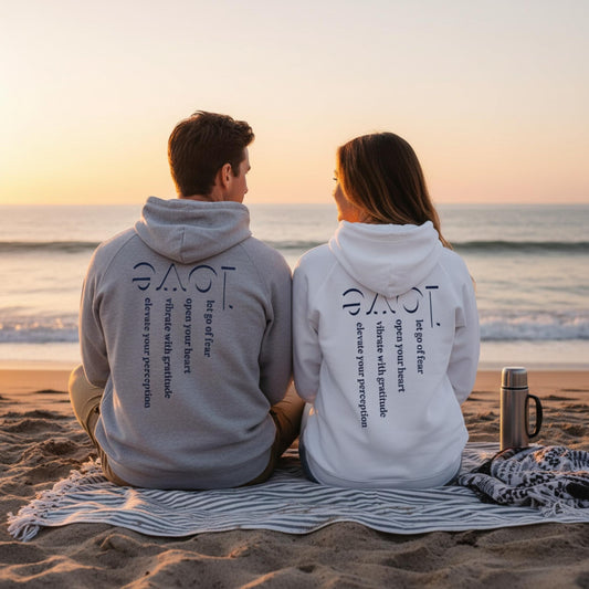 Two people sitting on a beach wearing hoodies with love design by Rose Fibonacci Designs, facing the ocean at sunset.